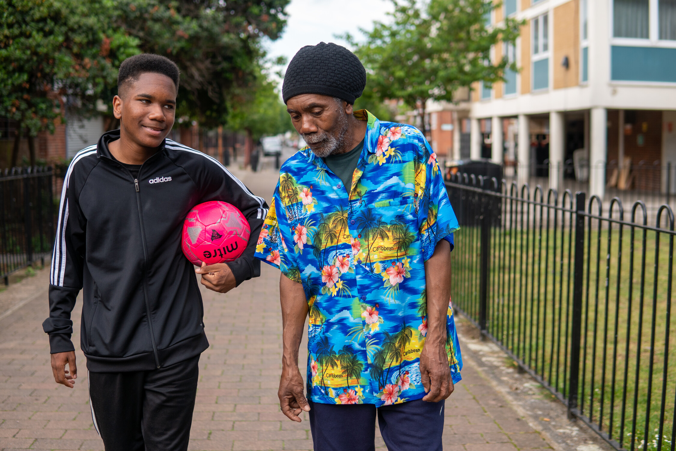 A boy and man in a tropical shirt walk down an urban street. The boy holds a ball.