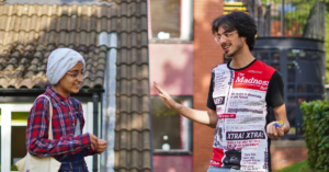 A young woman in headscarf stands outside a house and talks to a young man. They are both smiling and wearing glasses.