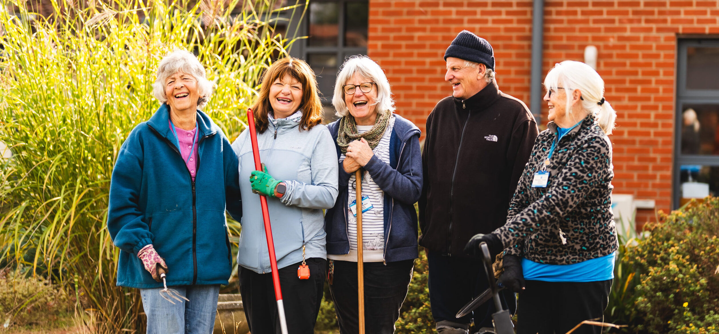 A group of 5 people in gardening gear outside. They are happy. There is a wheelbarrow in the front of the image.