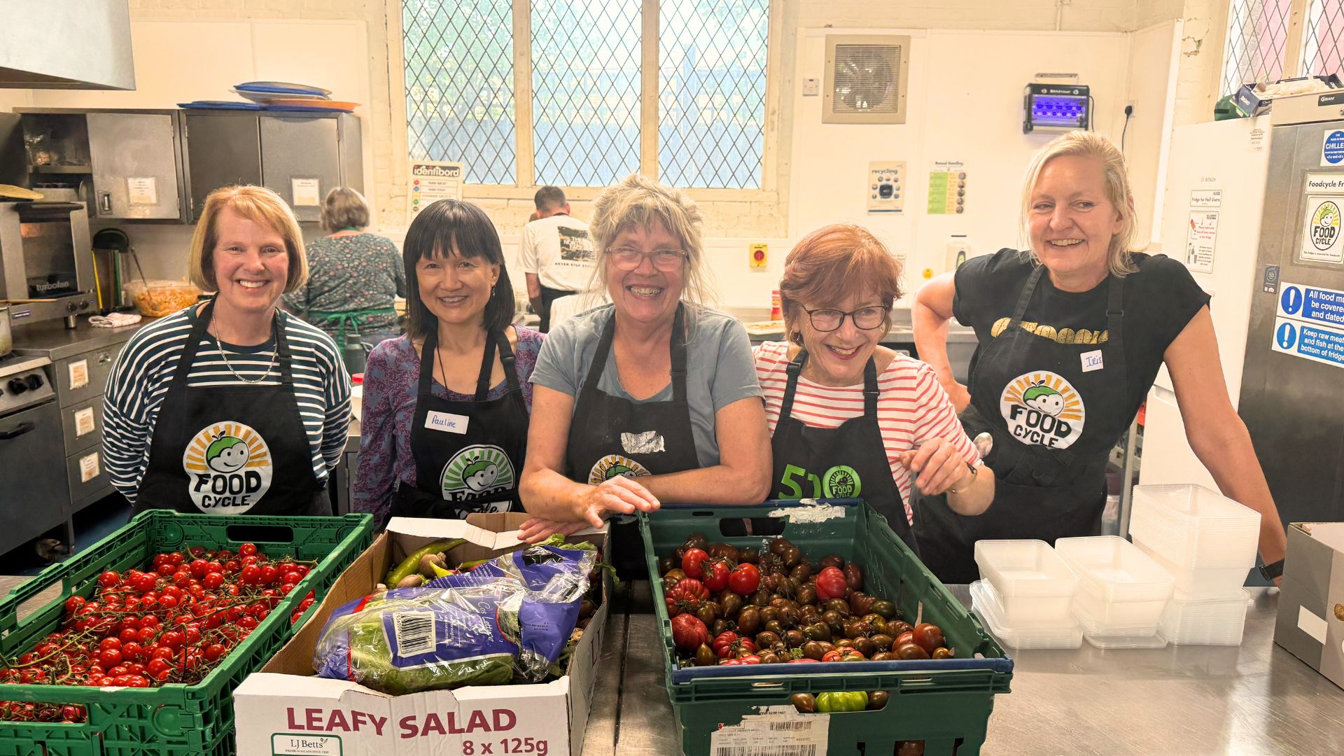 An image of 5 people in a kitchen. They are smiling wearing FoodCycle aprons