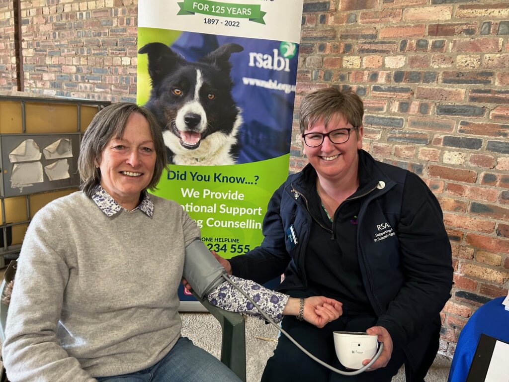A woman in a jumper has her blood pressure checked by a woman in a black top. They are smiling. There is a banner behind with a sheepdog on.