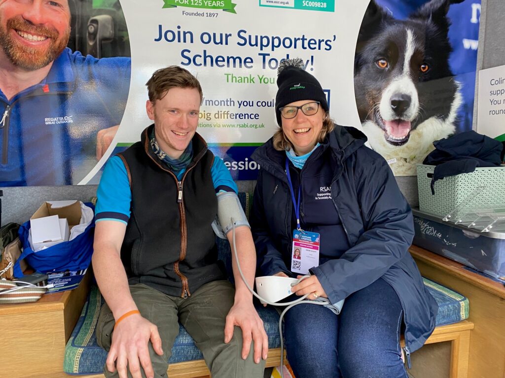 A man has his blood pressure checked by a woman in a bobble hate. They are smiling. There is a banner behind with a sheepdog on.