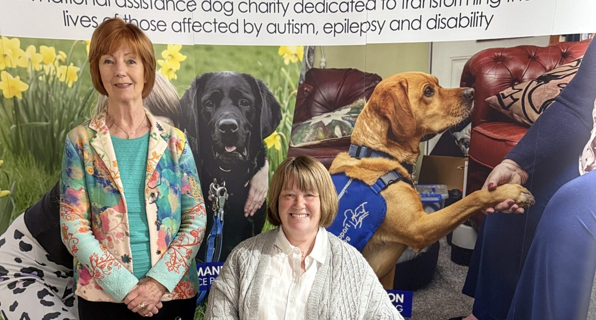Two women and a dog sit in front of a Support Dogs Banner