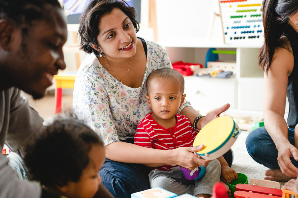 A woman with a baby on her lap hold a tamborine. A man sits on the edge with a baby on his lap