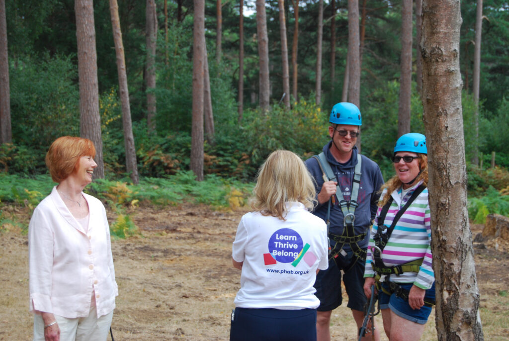 4 people in a wood. Two wear helmets and climbing gear. Two others are talking to them.